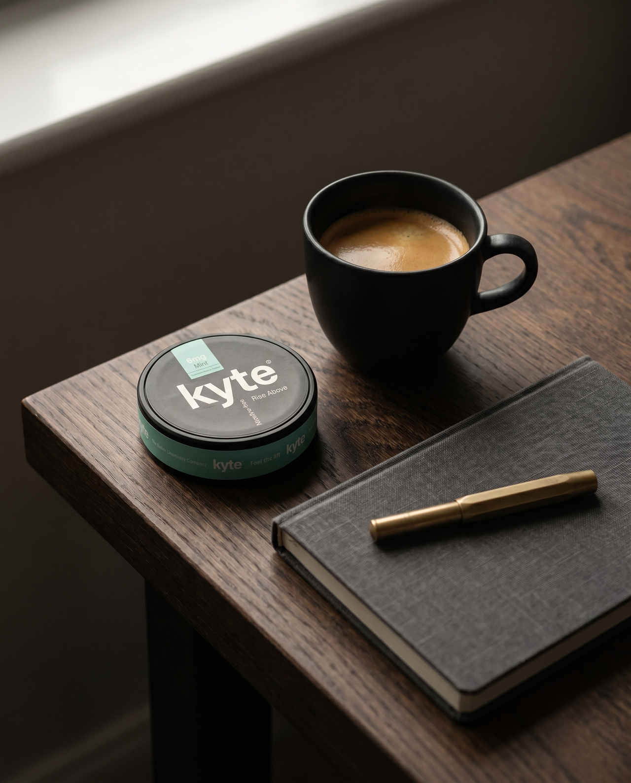 A Kyte tin beside a black espresso and a linen notebook on a dark oak desk